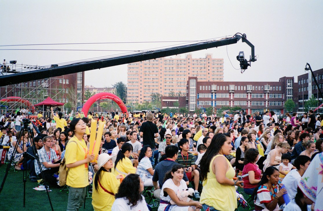 Large outdoor gathering of over 1,000 schoolchildren in an urban plaza with performers in yellow shirts and a red inflatable arch visible.