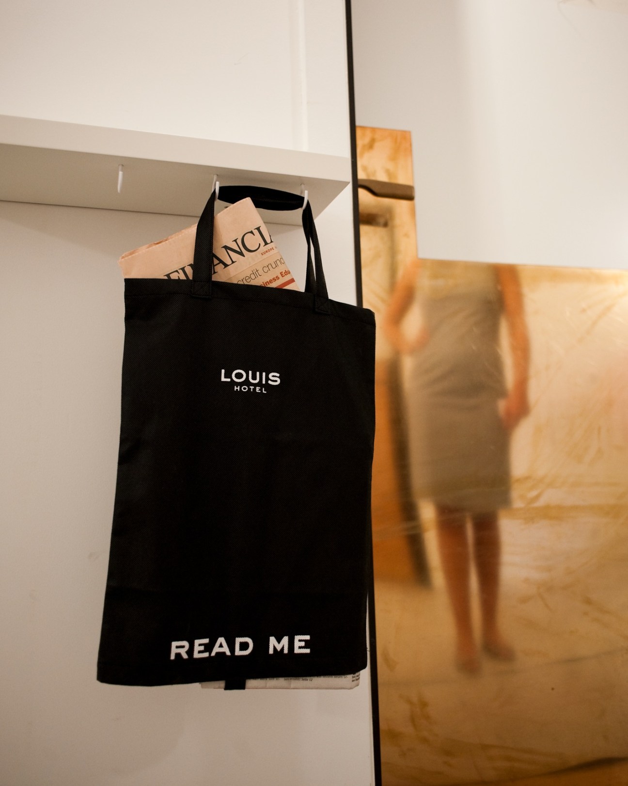 Black tote bag branded 'Louis Hotel' with 'Read Me' text hanging on white wall.