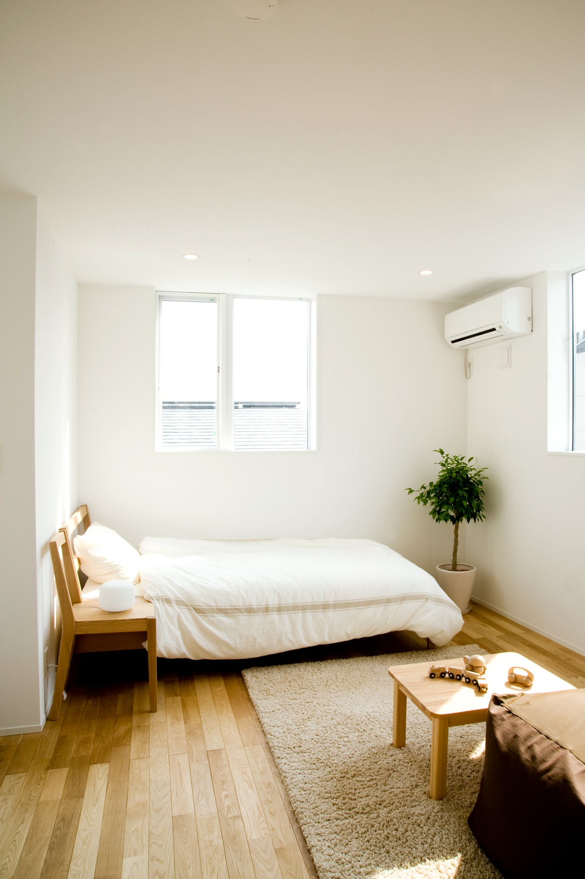 Minimalist bedroom interior with wooden bed frame, white bedding, potted plant, and natural light from windows.