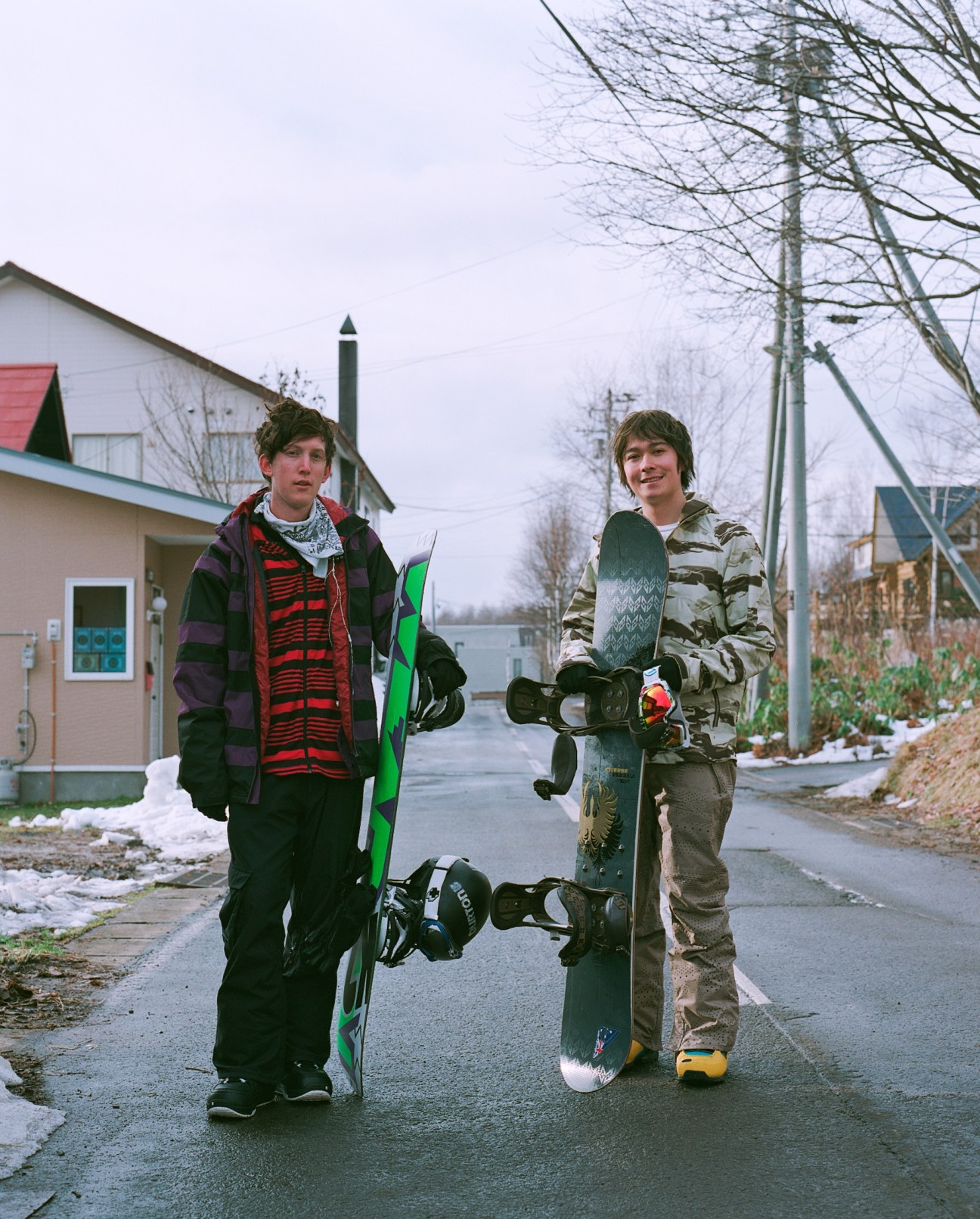 Two young snowboarders holding their boards stand on a residential street in Niseko.