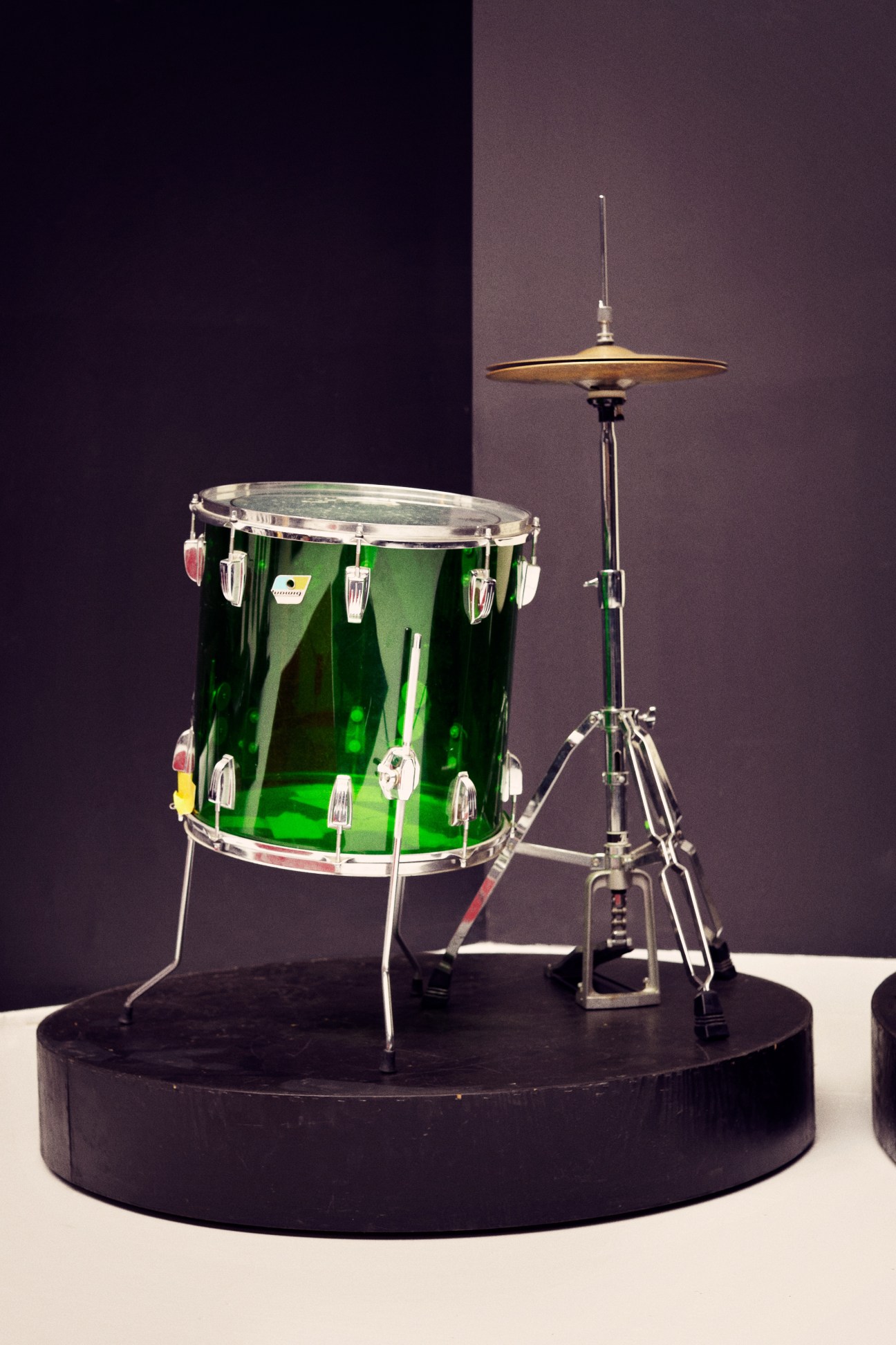 Green drum kit with floor tom and hi-hat cymbal on black platform against purple wall.