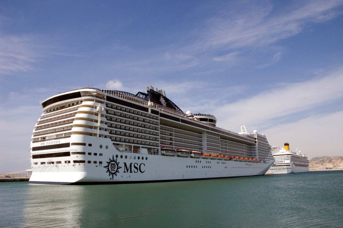The MSC Fantasia cruise ship docked in port, showing its 18-storey white hull with brown and tan striping.