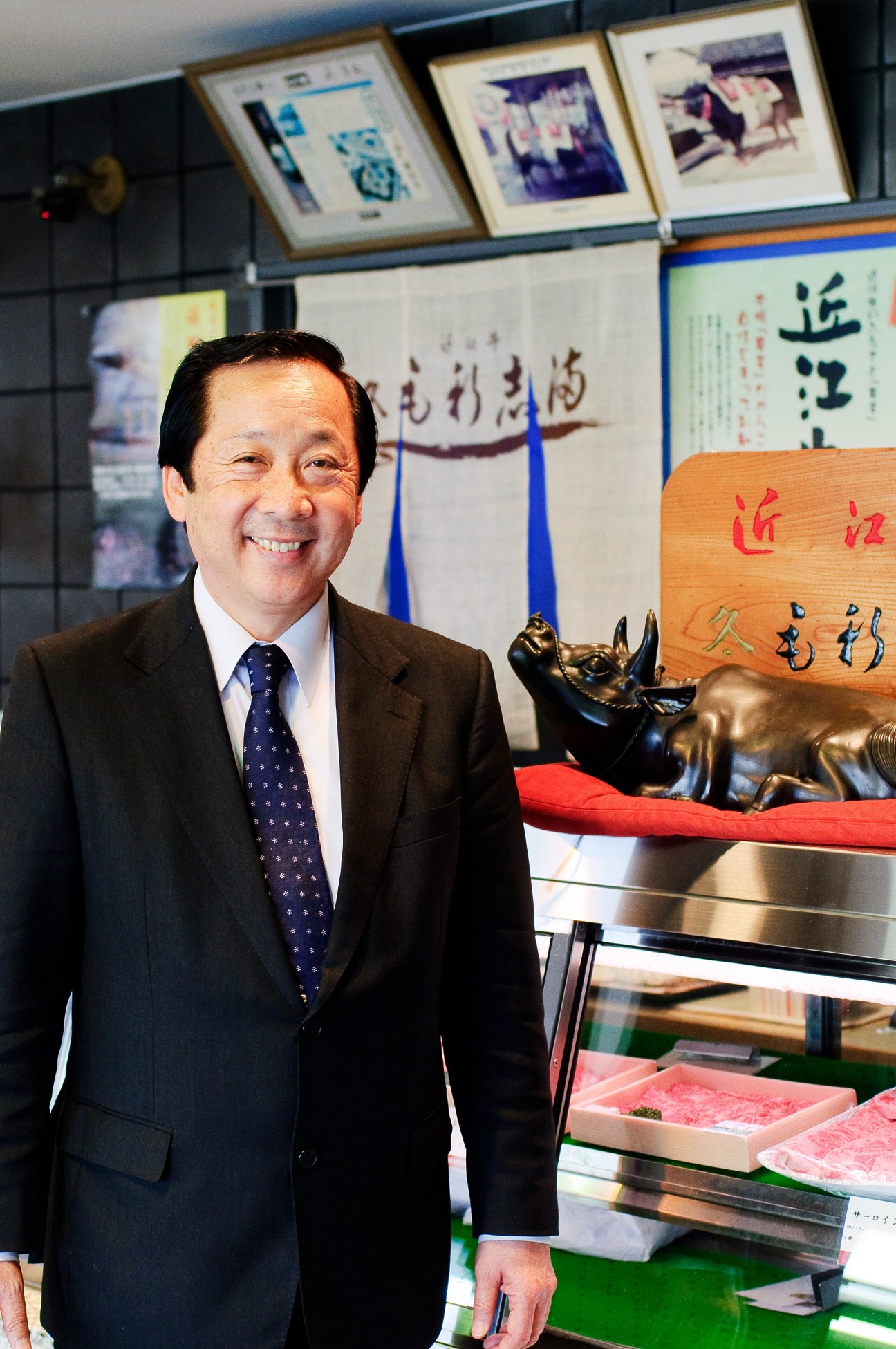 Tokuo Morishima smiles at Morishima restaurant, with Japanese calligraphy and a bronze cow sculpture visible behind him.