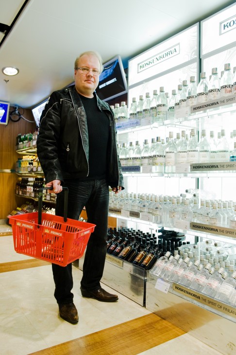 Man with shopping basket stands in front of shelves stocked with Koskenkorva vodka bottles in ship's duty-free shop.