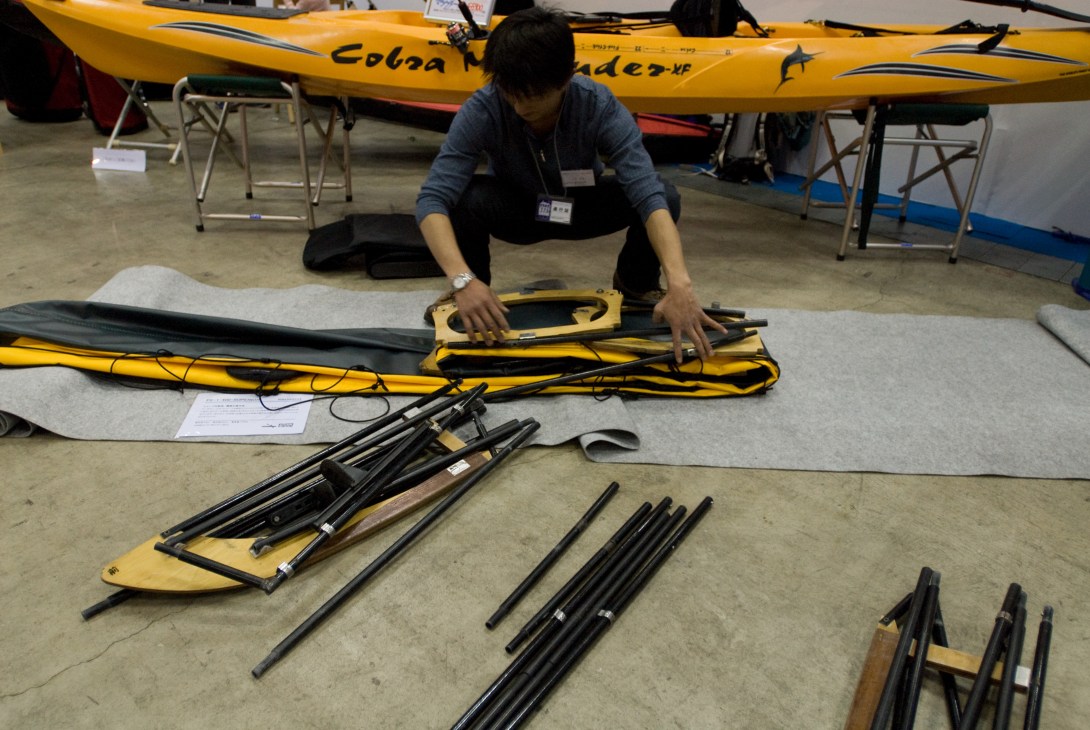 A woman demonstrates a yellow folding kayak with its components disassembled at a boat show display.