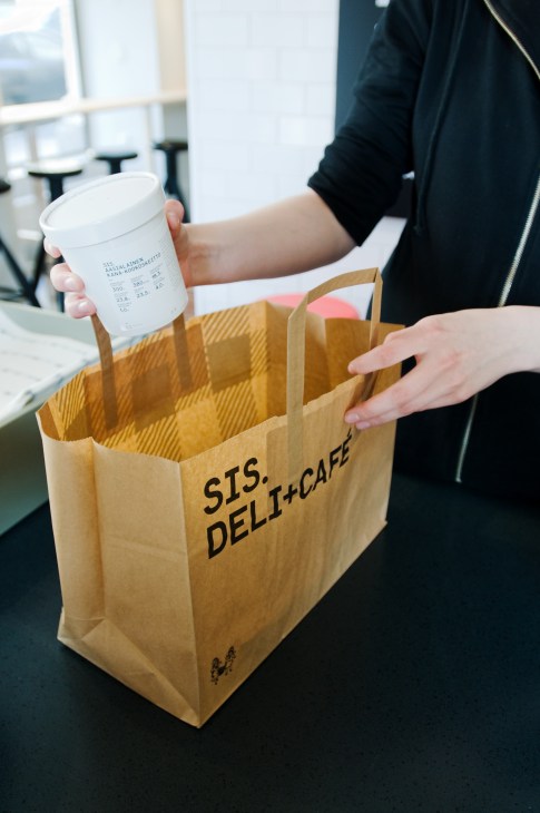 Person holding a Sis deli+café paper bag and white coffee cup in a bright cafe interior.