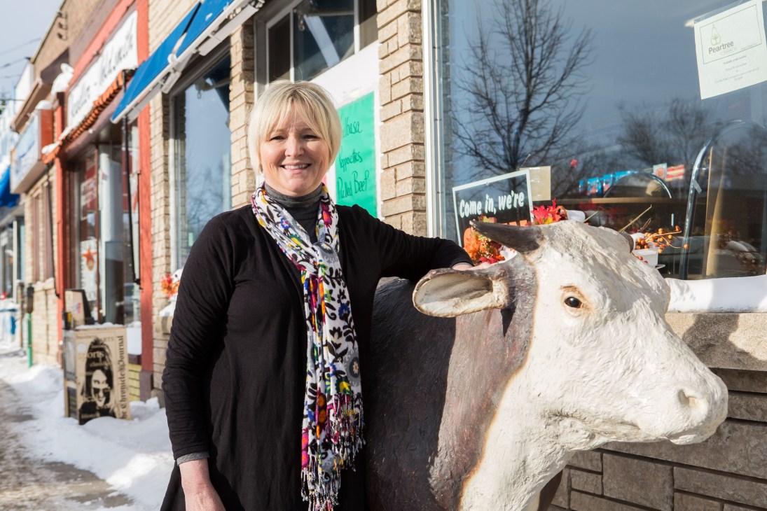 Cindy Salo stands outside Bay Meats Butcher Shop next to a carved wooden cow sculpture on a snowy Thunder Bay street.