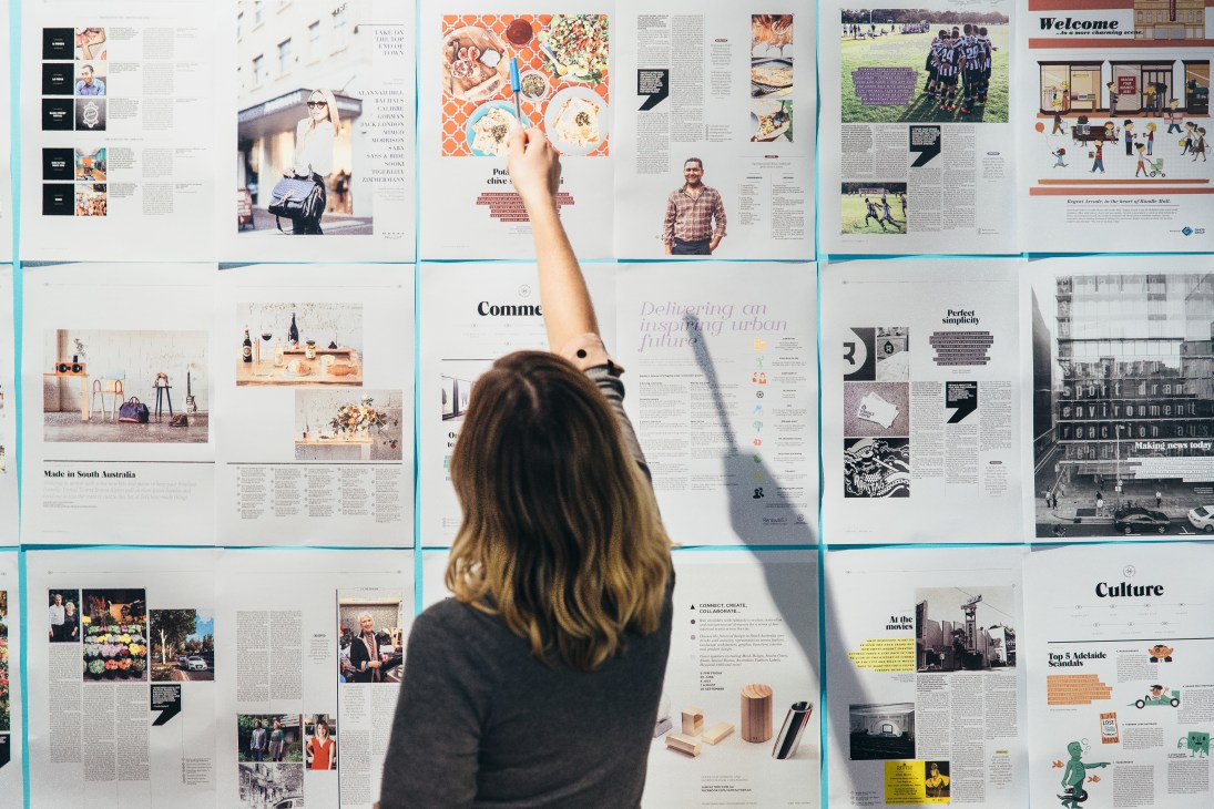 Editor Farrin Foster reaches toward magazine spreads displayed on a wall, reviewing layout and design.