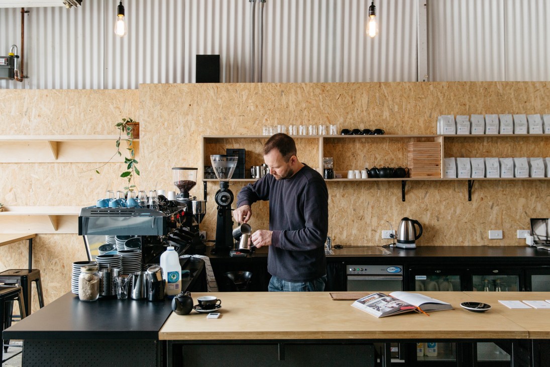 Barista at Tell Henry café pouring milk into coffee while standing behind counter with espresso machine and shelving.