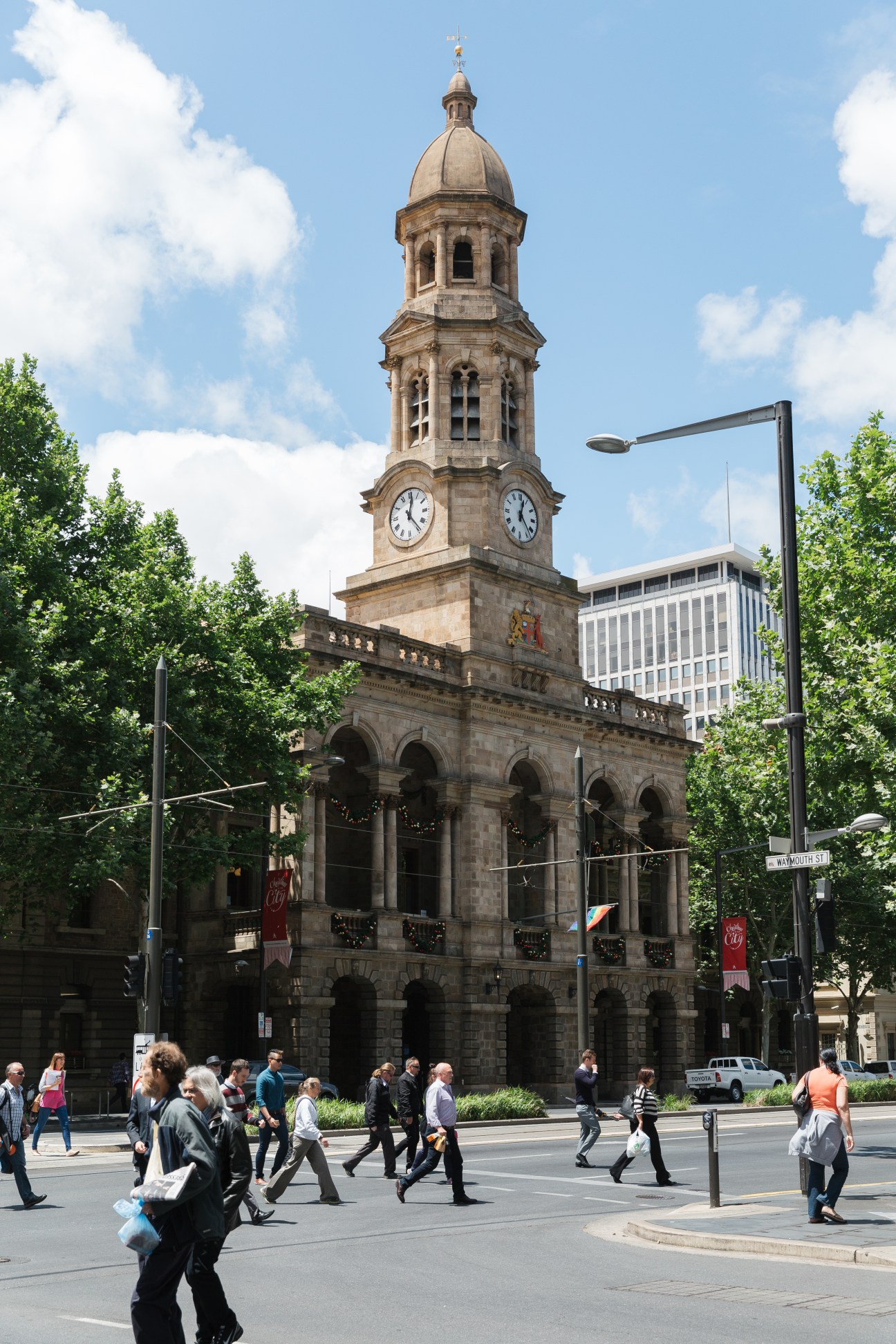 Adelaide Town Hall's historic stone clock tower with dome amid surrounding modern buildings and pedestrians.