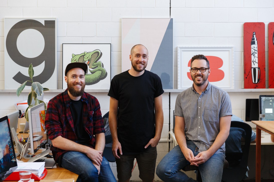 Three men posing in a creative office space with design artwork on the wall behind them.
