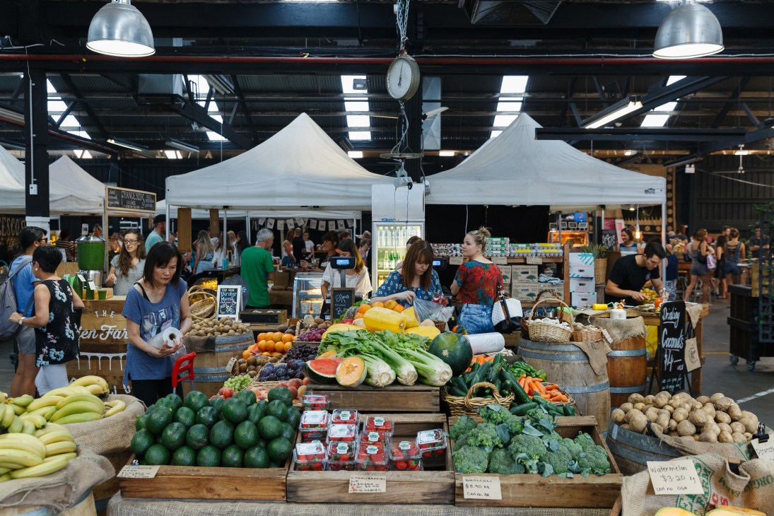 Bustling farmers market with fresh produce displays under white tents and industrial ceiling.