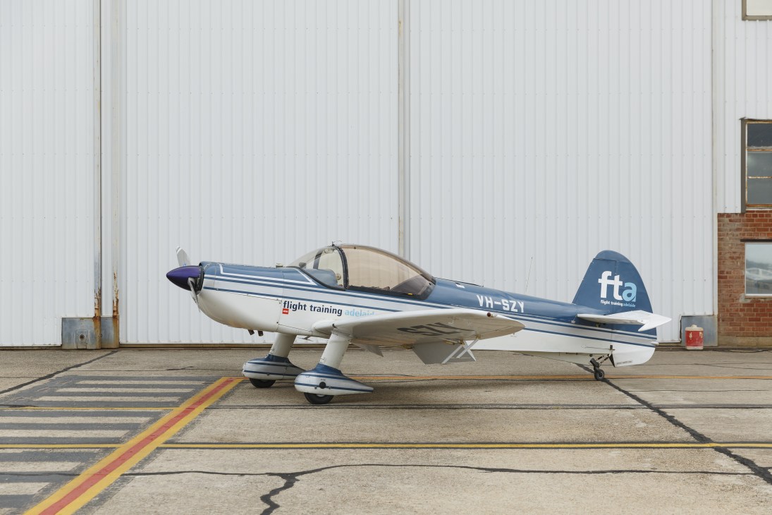 A blue and white Mudry CAP 10 aircraft parked on the tarmac outside a hangar at Flight Training Adelaide.