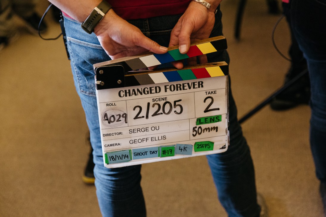 A film clapperboard for 'Changed Forever' showing scene 2/205, take 2, held by a crew member on set.