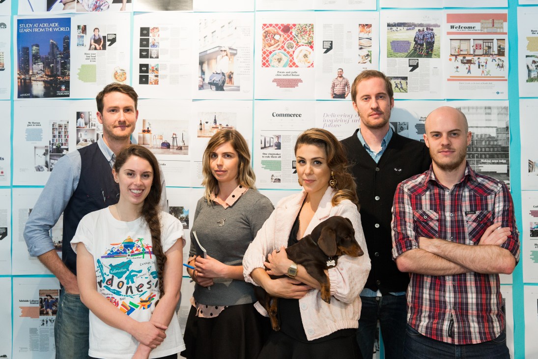 The CityMag team of six people posing in front of a wall of magazine layouts and design work.
