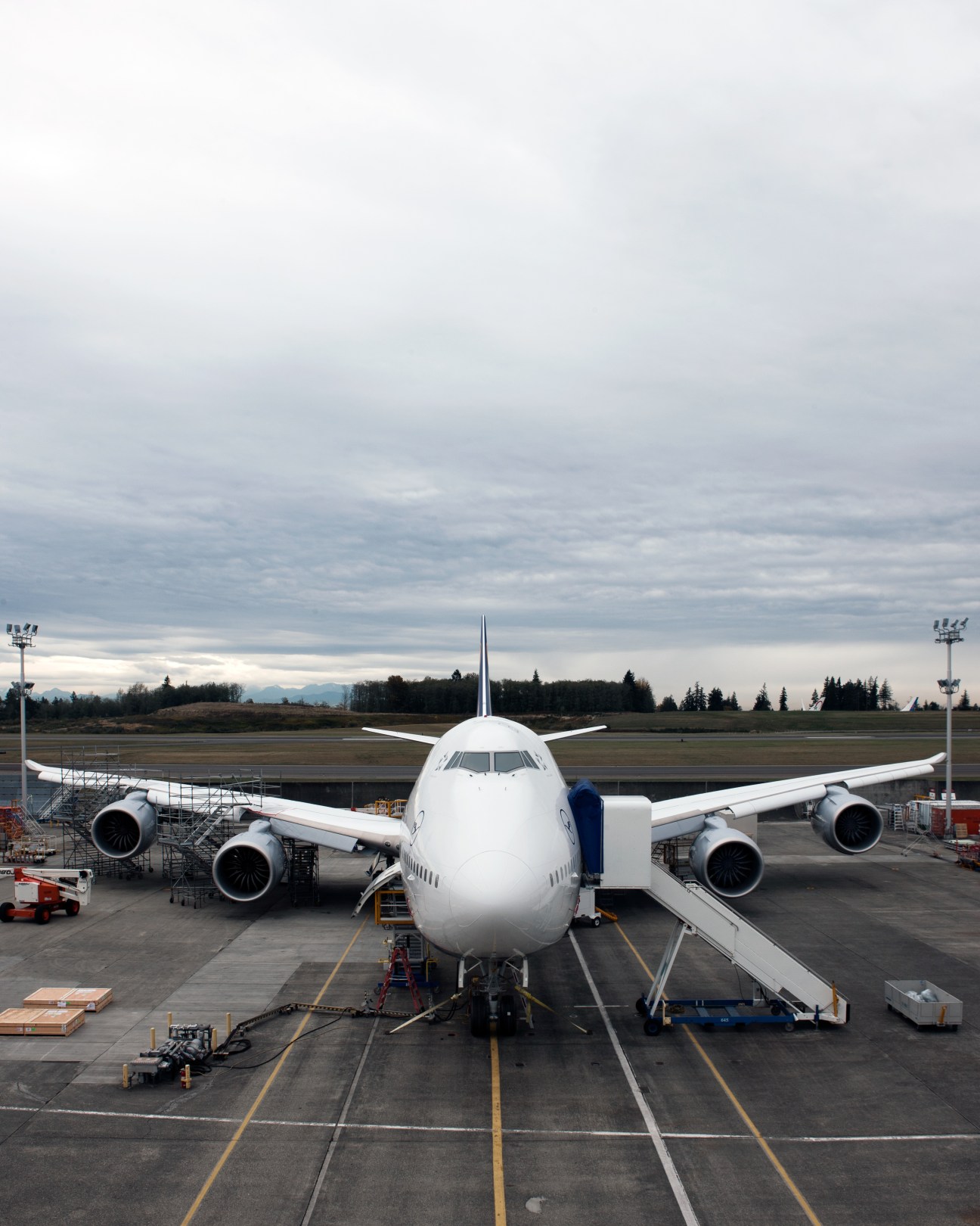 Boeing 747-8 aircraft parked at airport apron with ground equipment and evergreen trees in background.