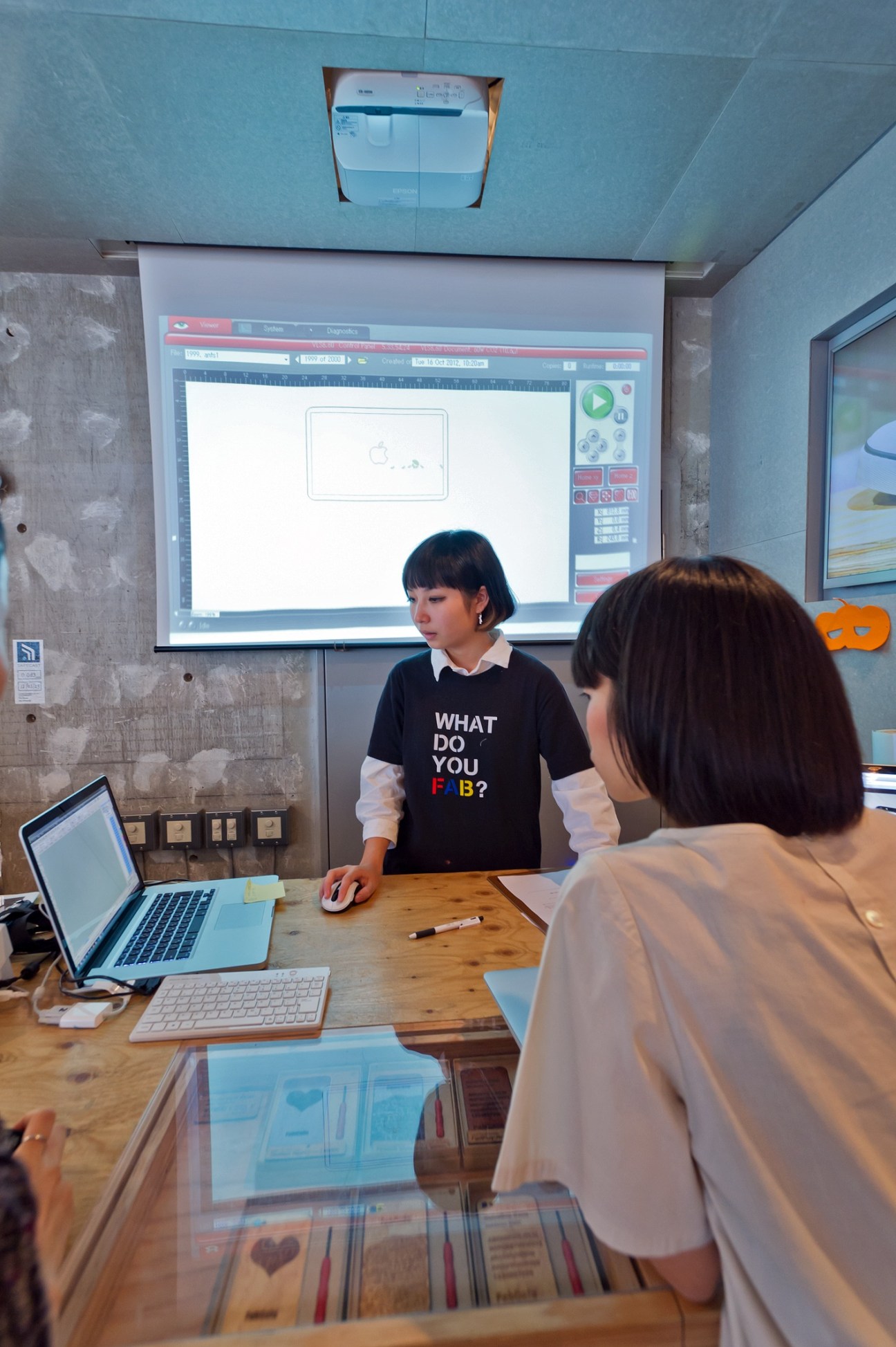 Woman in black sweater standing at wooden desk with laptop, facing another person, with digital design displayed on wall screen.