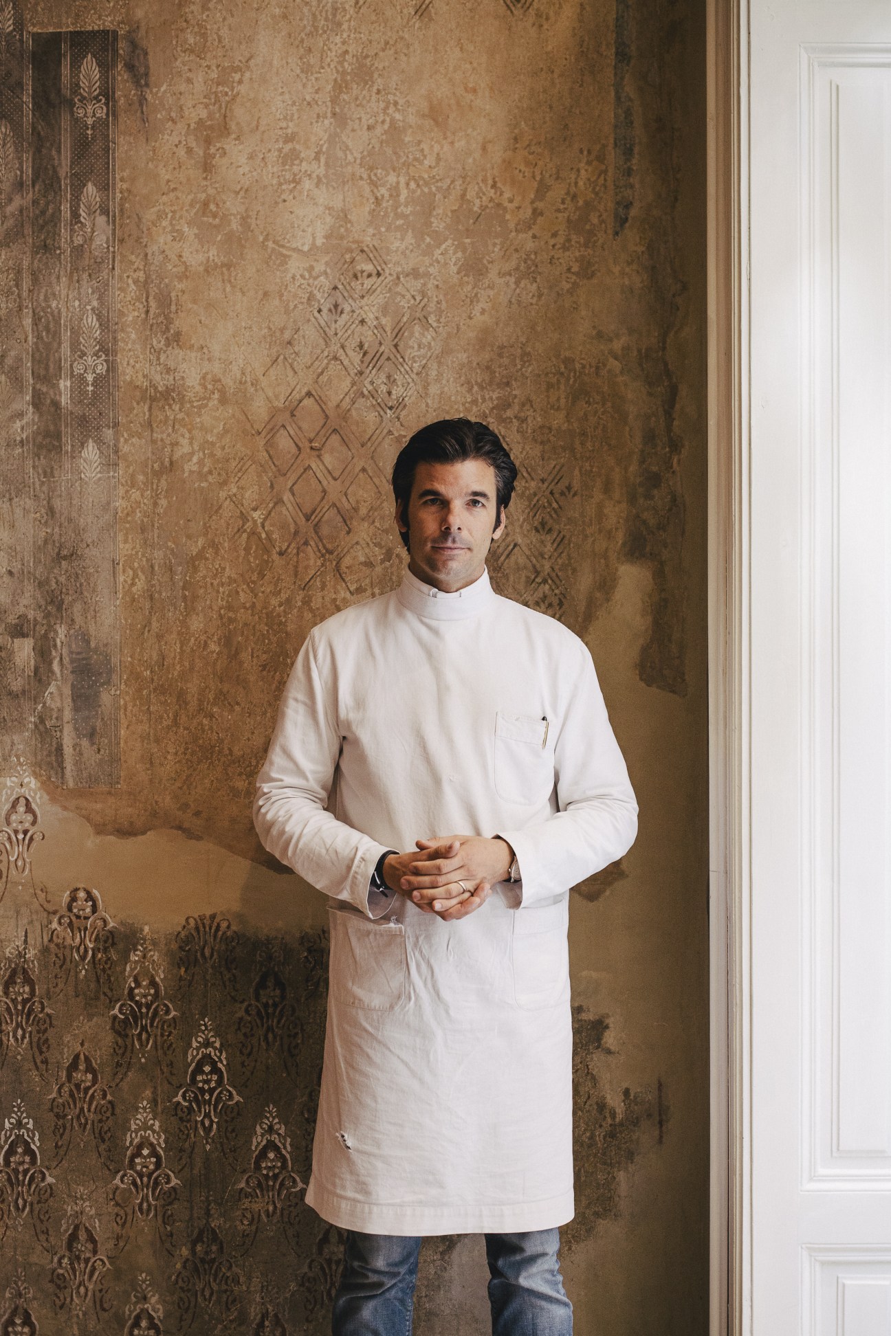 Man in white linen tunic standing against ornate vintage wallpaper in Rudolf Scheer & Söhne shop
