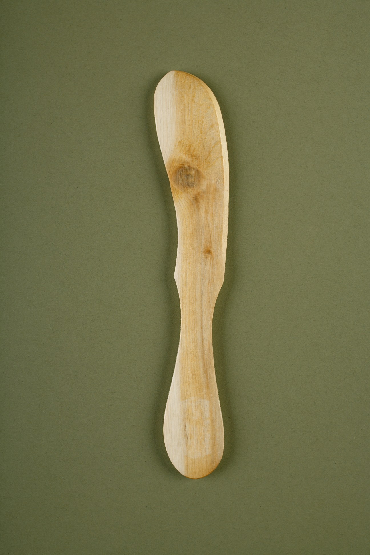 A wooden butter knife with curved edges and a flat spreading surface against a sage green background.