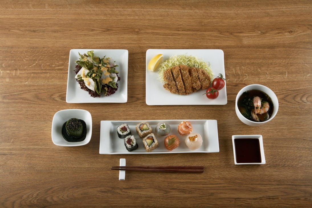 Japanese dishes including sushi, tonkatsu, salad and soup arranged on white plates with chopsticks and sauces on wooden table.