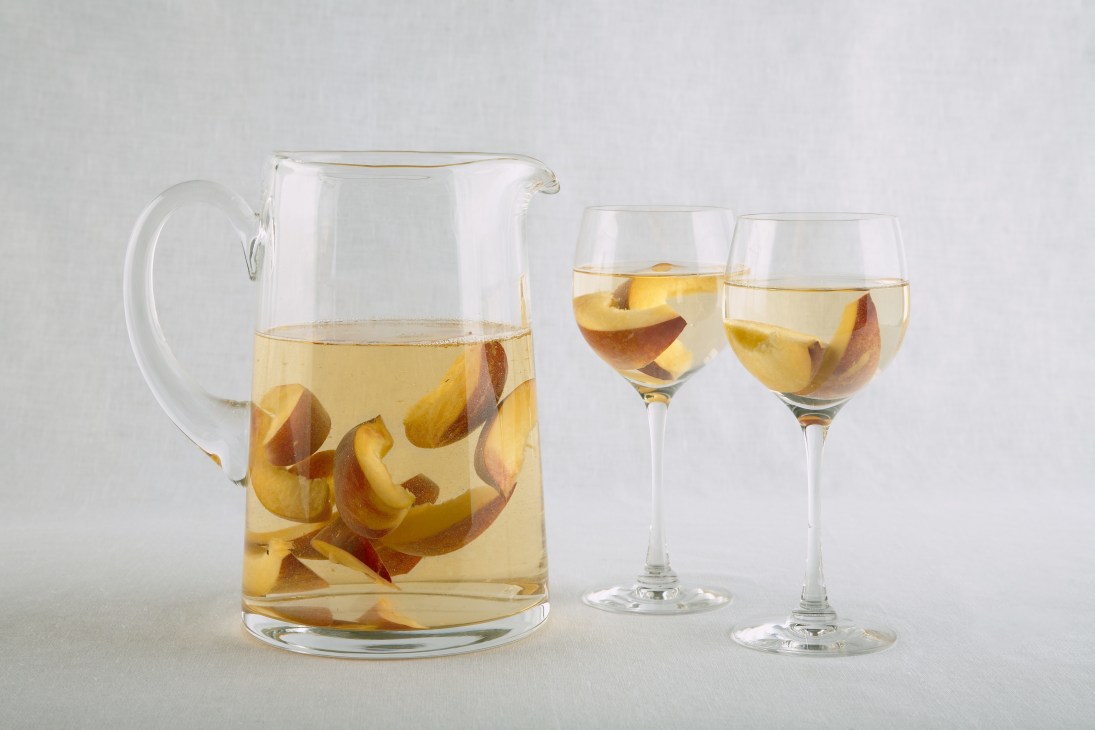 A glass pitcher and two wine glasses filled with white wine and fresh peach slices.