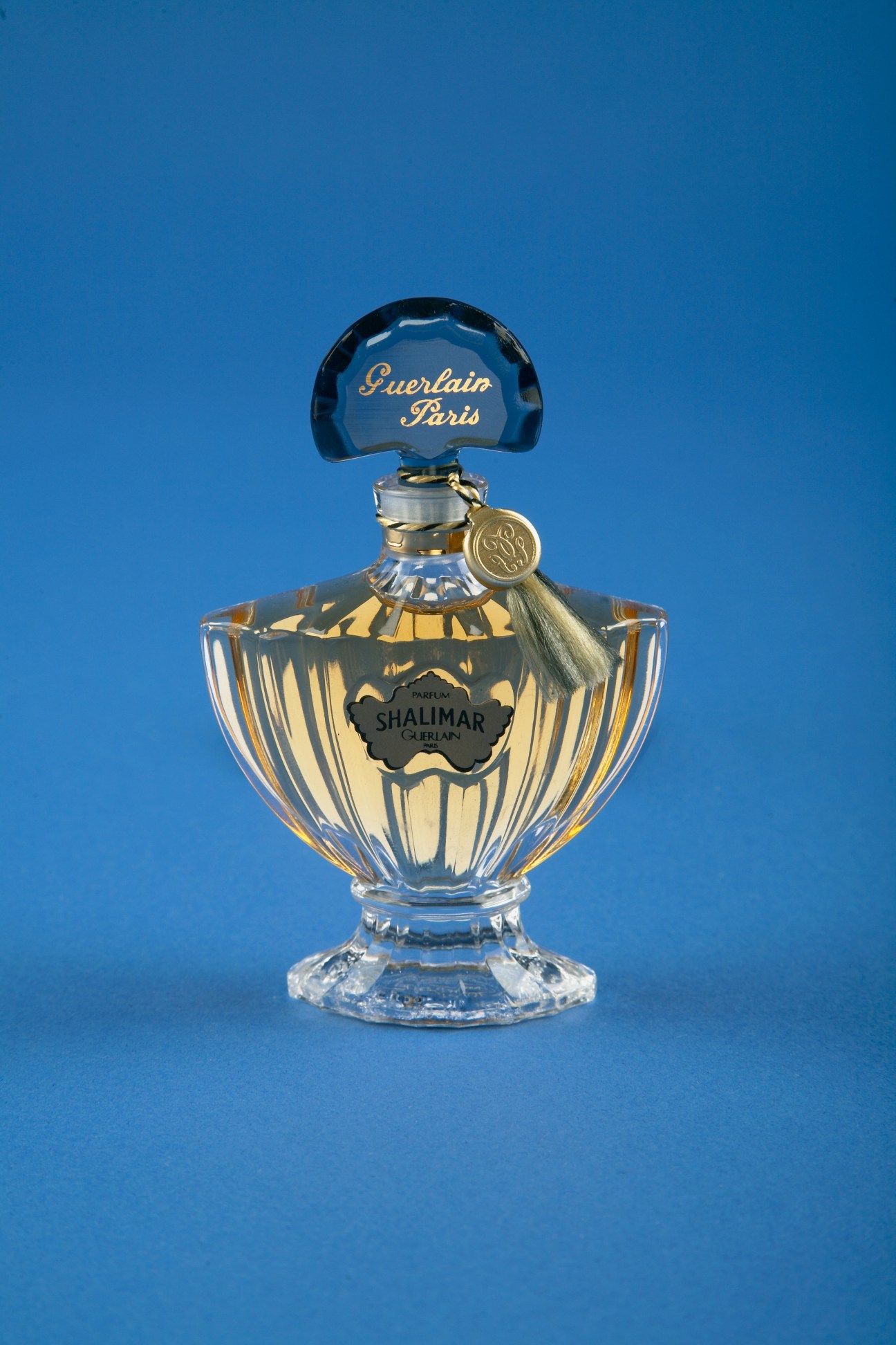Guerlain Shalimar perfume bottle with blue stopper and tassel against bright blue background.