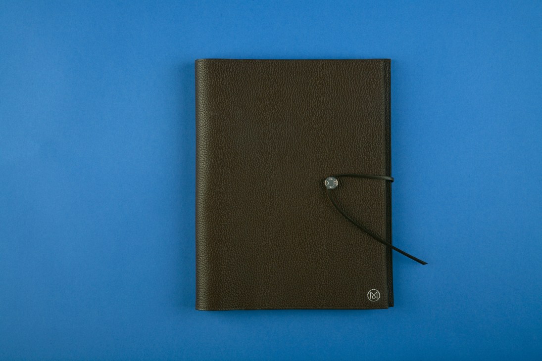 Olive-green leather Hermès journal with Monocle monogram and black ribbon bookmark on blue background.