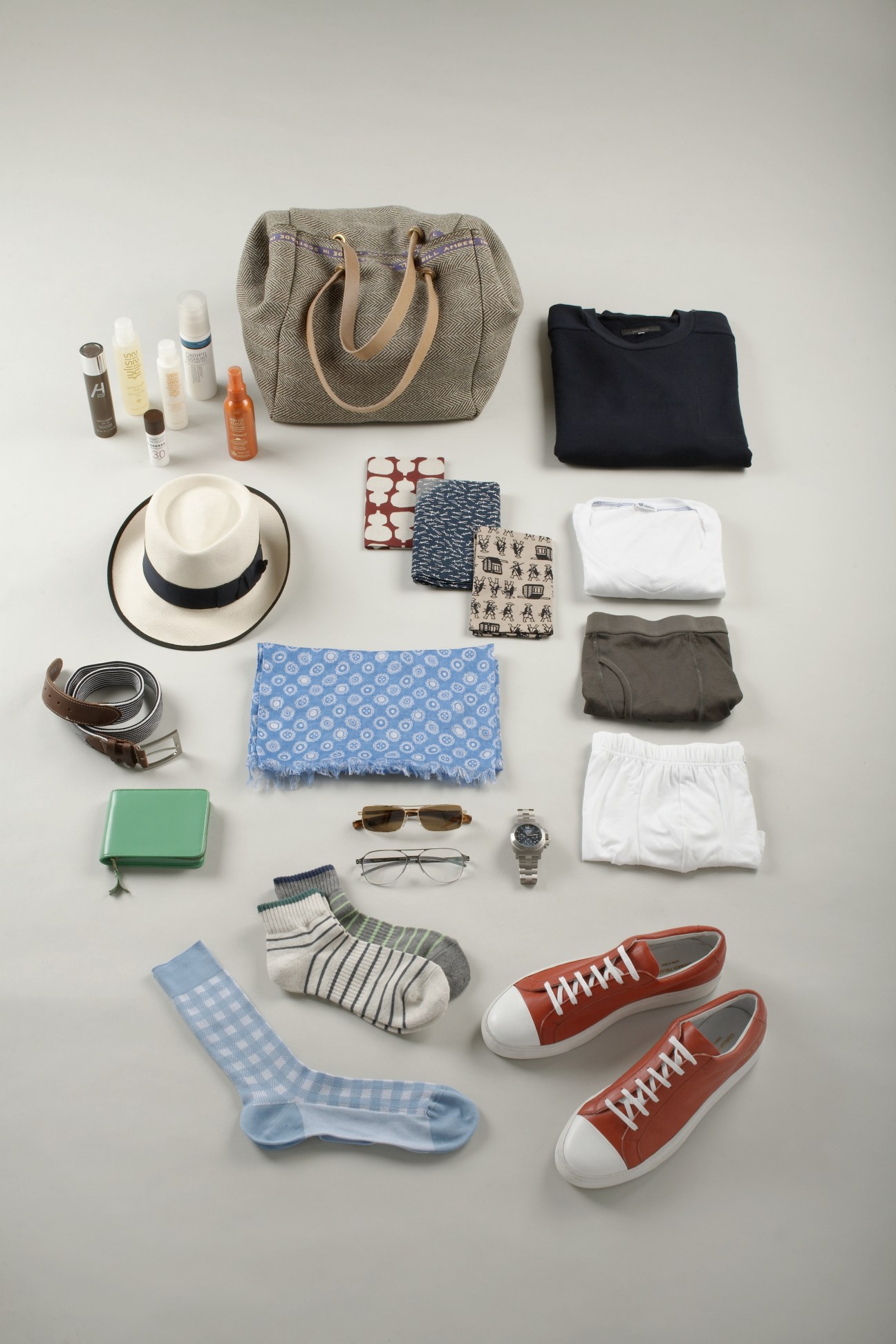 Flat lay of men's accessories including canvas bag, hat, belt, sunglasses, watch, socks, shoes, scarf and grooming products