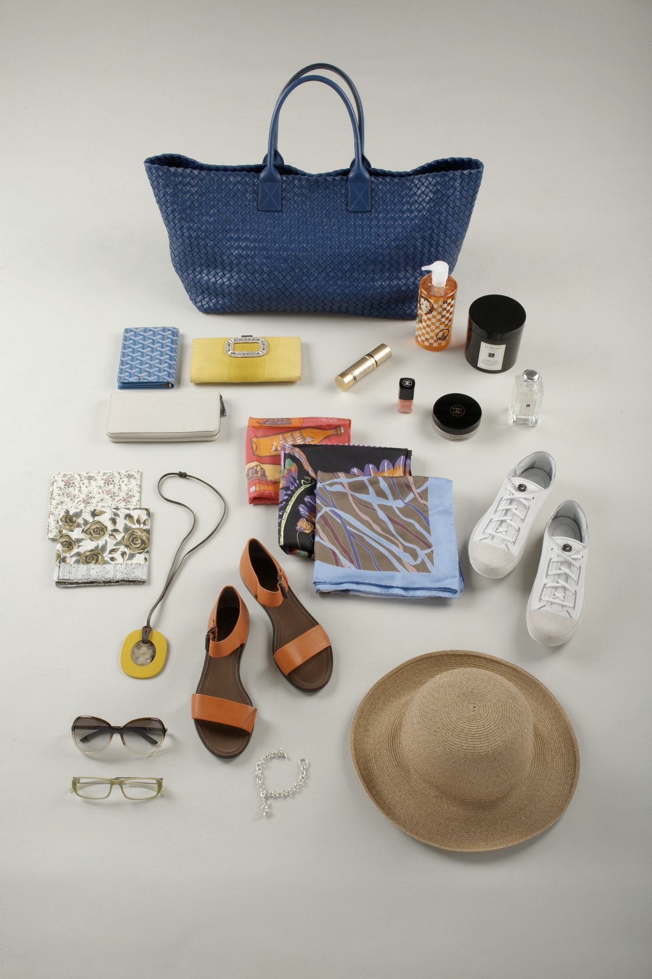 Flat lay of women's accessories including a blue woven tote bag, yellow wallet, orange sandals, sunglasses, scarves, beauty products, sneakers and a straw hat.