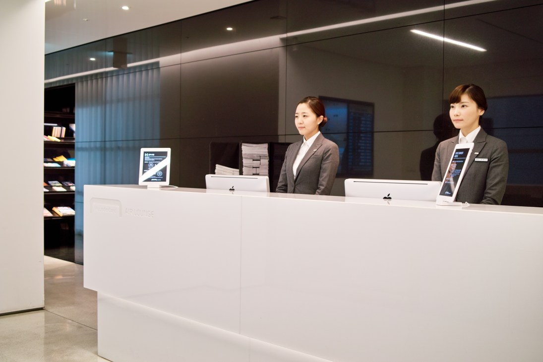 Two female staff members stand behind a white reception desk in a modern minimalist interior with dark walls and ambient lighting.