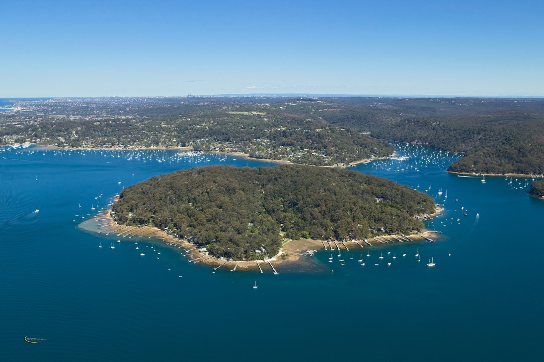 Aerial view of Scotland Island, a forested island surrounded by blue water with moored sailboats and nearby coastal suburbs.
