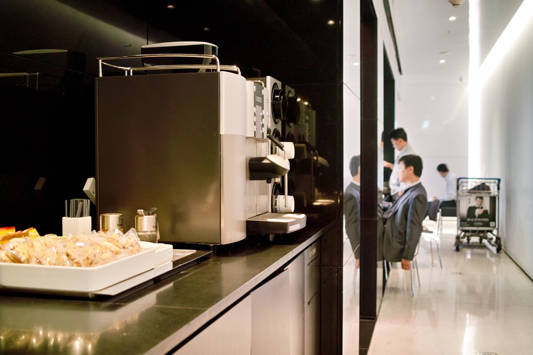 Modern minimalist lounge interior with sleek espresso machine and seating area, customers visible in background.