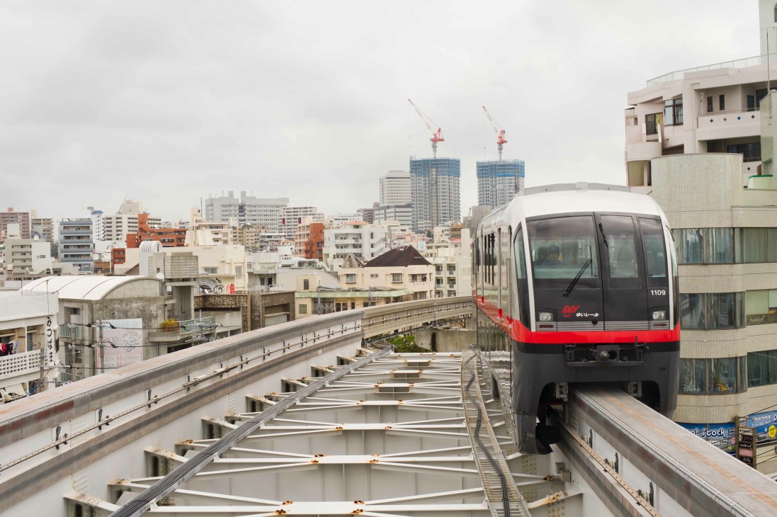 A red and gray elevated train with Japanese characters on its side travels on tracks above an urban Japanese cityscape with residential and commercial buildings.