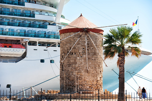 Large cruise ship docked next to a historic stone windmill with palm trees in Mandraki Harbour, Rhodes.