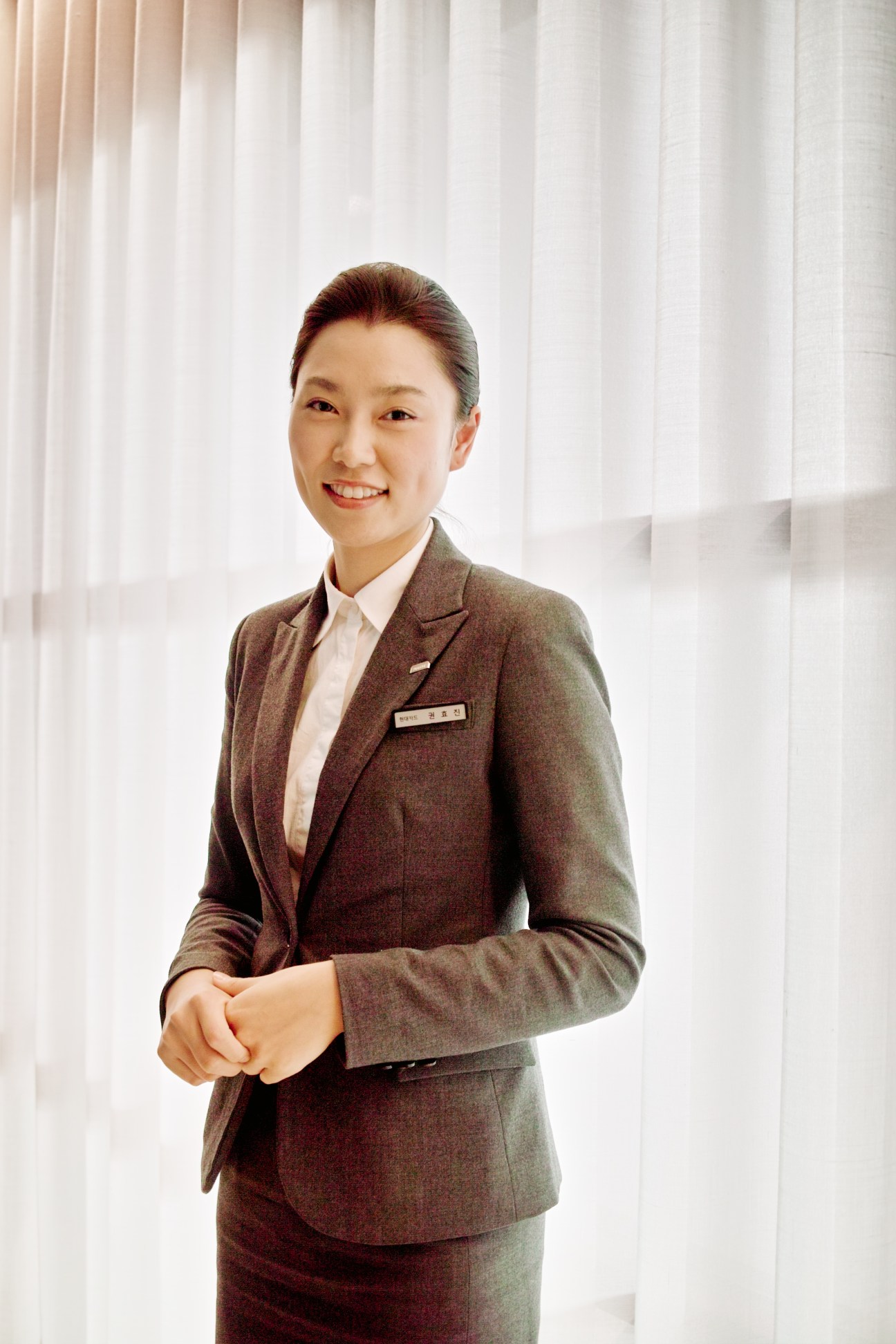 Female hotel staff member in brown blazer and white shirt smiling at camera against white curtained background