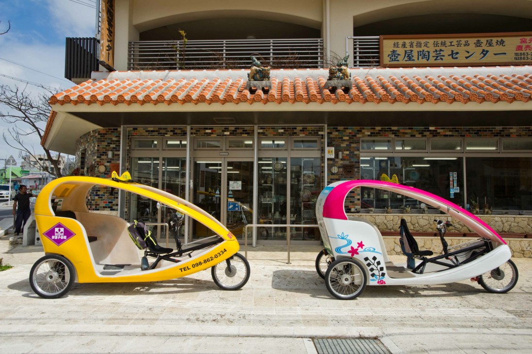Two colorful electric rickshaws parked in front of a traditional Japanese building with orange tile roof in Naha.