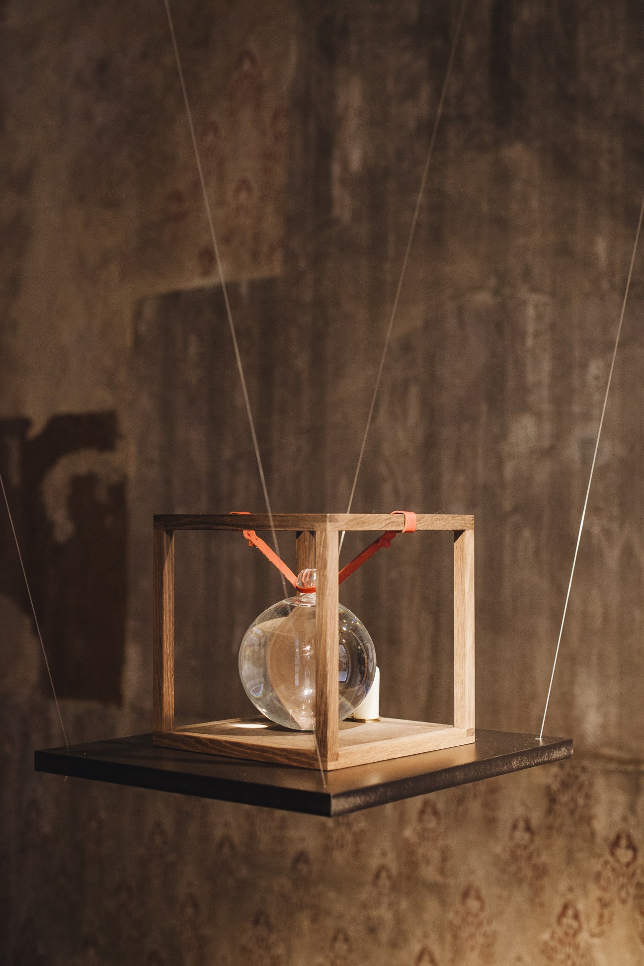 Minimalist wooden cube clock with glass sphere and orange and red hands suspended by wires.