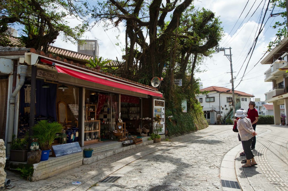 Street scene in Naha showing a traditional shop with red awning beneath large trees, with pedestrians and buildings visible.