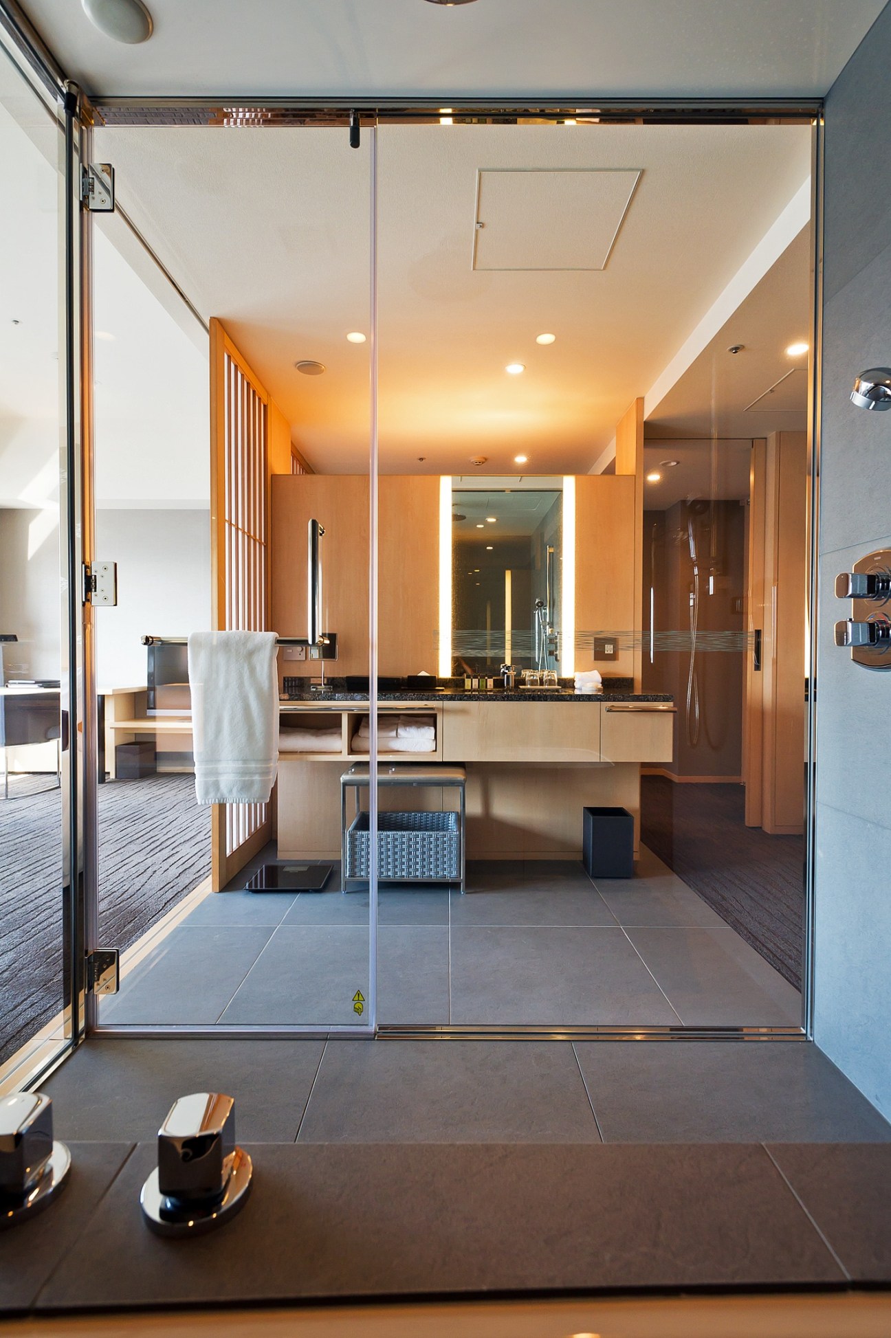 Modern hotel bathroom with wooden vanity, illuminated mirror, glass partition, and spa facilities visible.