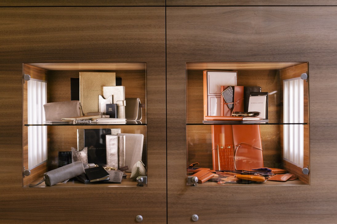 Wooden display cabinet with glass doors showing leather goods and accessories in neutral and orange tones.