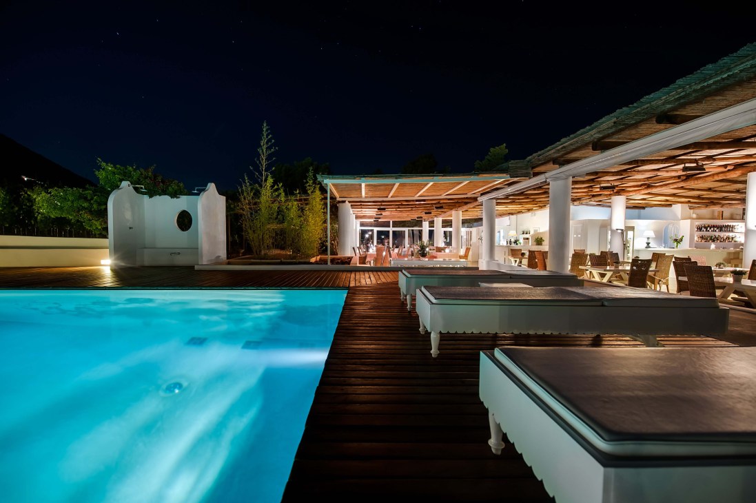 Night view of Capofaro resort's illuminated pool, wooden deck, and whitewashed buildings on Salina island.