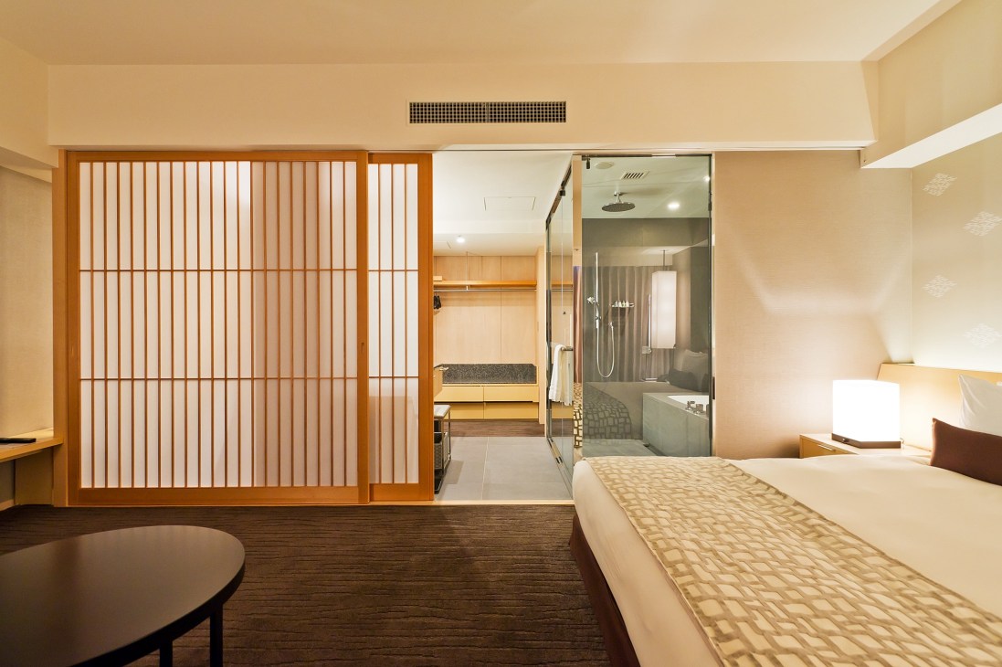 Modern hotel room with wooden sliding door, bed, and open bathroom with glass shower enclosure.