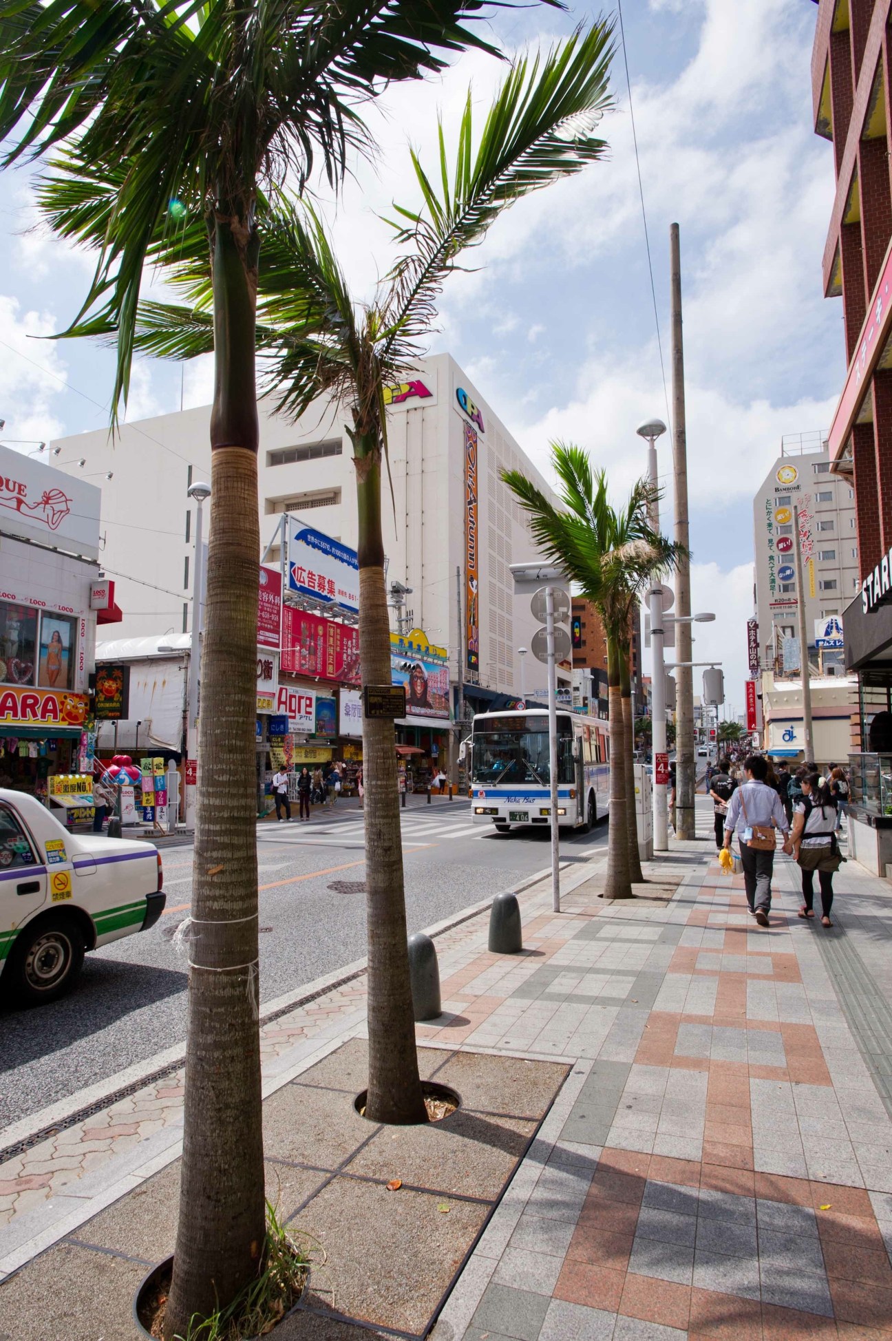 Urban street scene in Naha with palm trees, pedestrians, shops and buildings under blue sky.