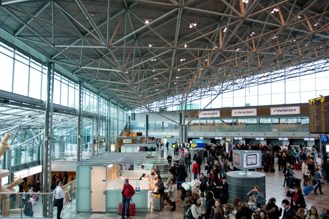 Helsinki airport terminal with steel lattice ceiling, glass walls, and crowds of travelers at check-in counters.