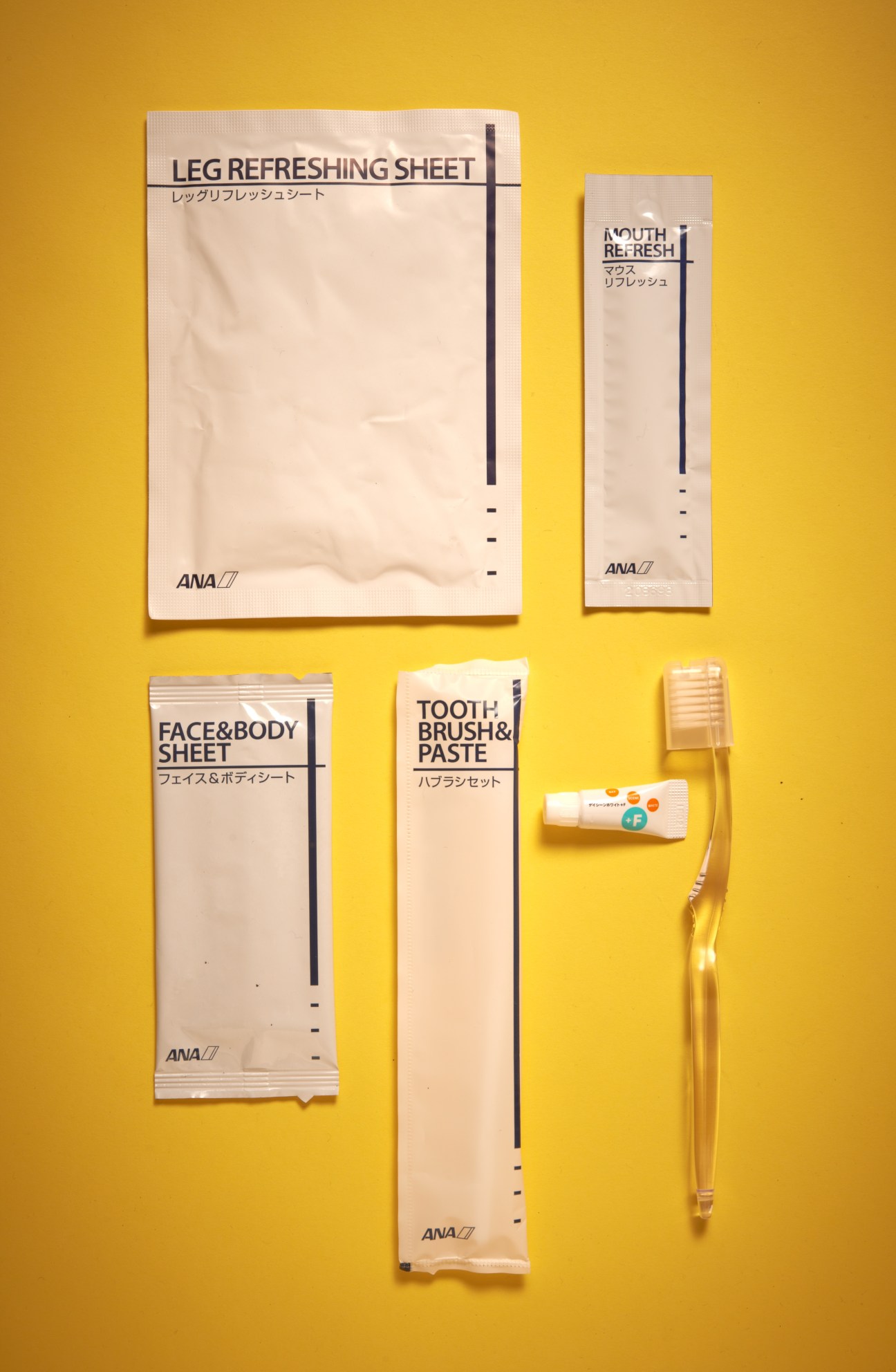 ANA airline amenity kit with leg refreshing sheet, mouth refresh, face and body sheet, toothbrush and paste on yellow background.