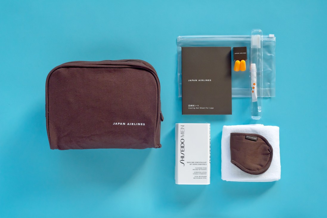 Japan Airlines amenity kit displayed on turquoise background with maroon pouch, Shiseido products, and accessories.