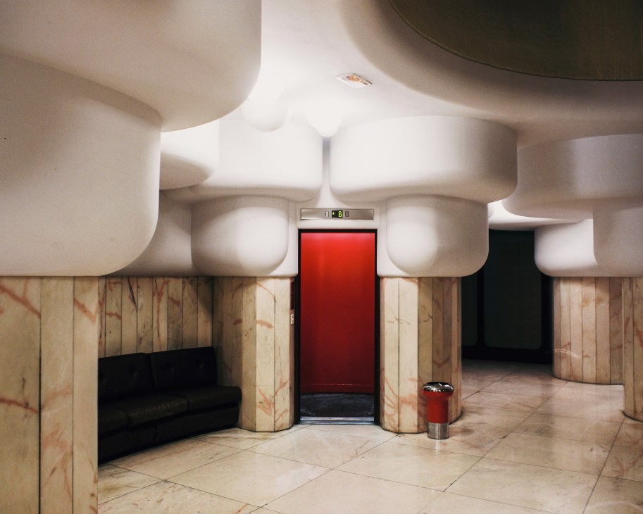 Torres Blancas lobby with curved white ceiling, marble walls, wooden pillars, red elevator door and red trash bin.