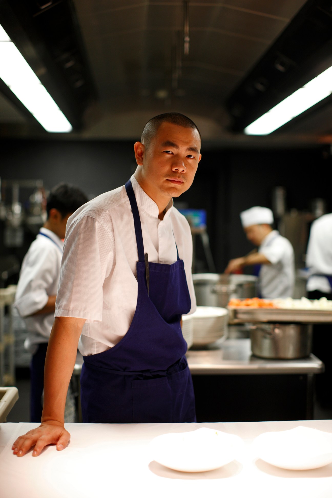 Chef in white shirt and blue apron stands in professional kitchen with staff working in background.