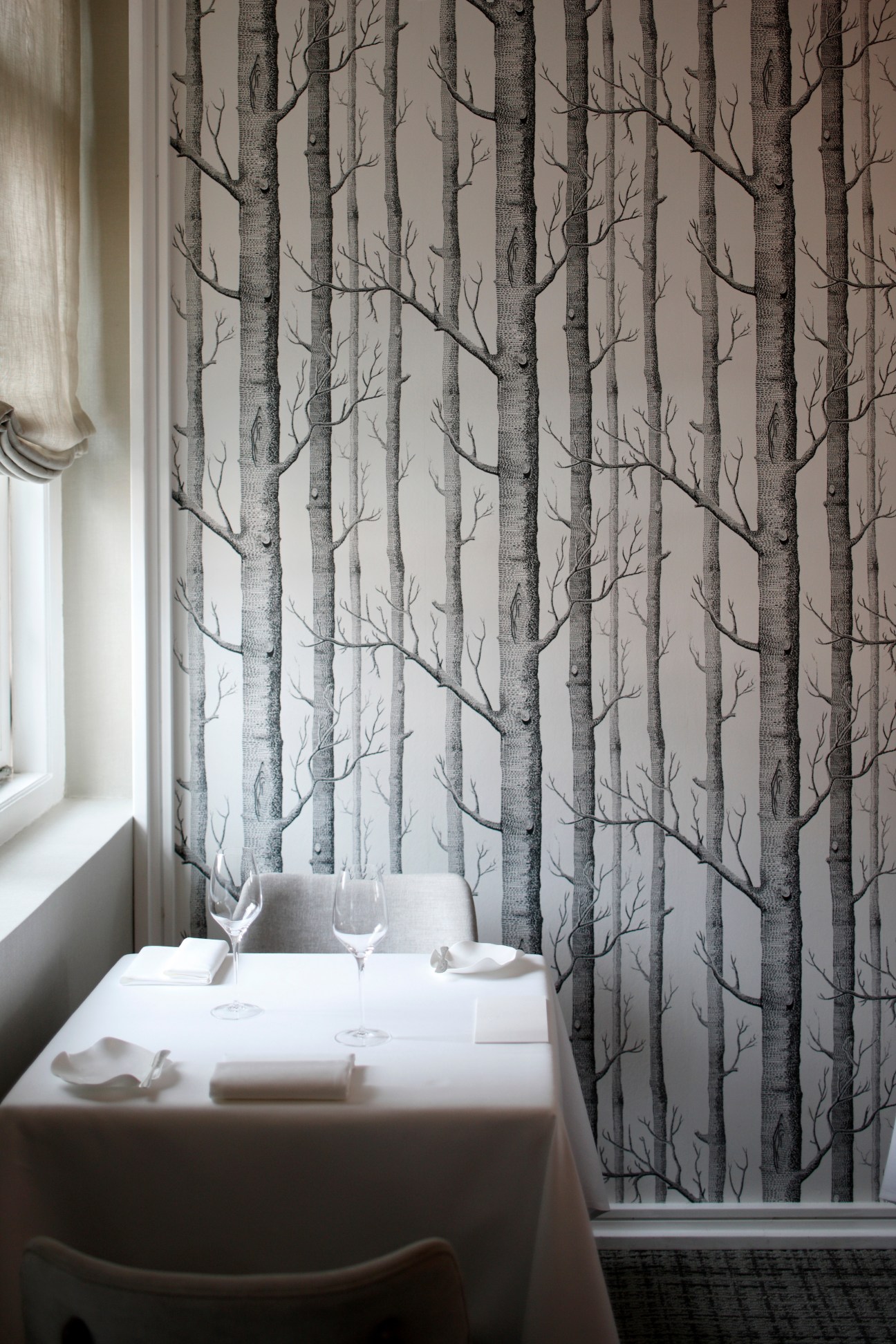 Restaurant dining room with forest wallpaper, set table, wine glasses and window with white curtain.