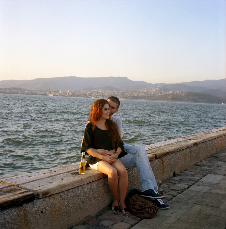 A couple sits together on a waterfront pier at sunset overlooking Izmir's bay and mountains.
